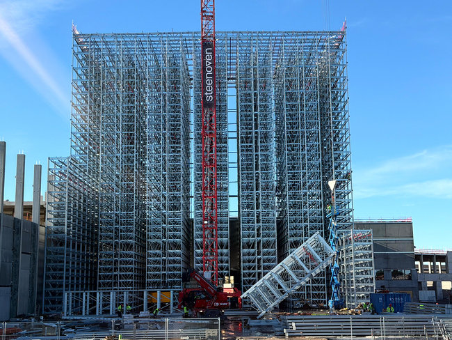 Les travaux de construction du b&acirc;timent-silo avancent &agrave; grands pas : une grande grue est en train de hisser l'une des sections de rayonnage pr&eacute;assembl&eacute;es &agrave; sa hauteur d'installation de 45 m&egrave;tres.<br>
            Cr&eacute;dit photo : Dematic